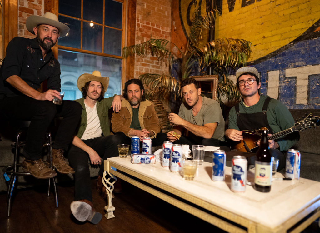 Group of guys sitting around a coffeetable covered with beer cans