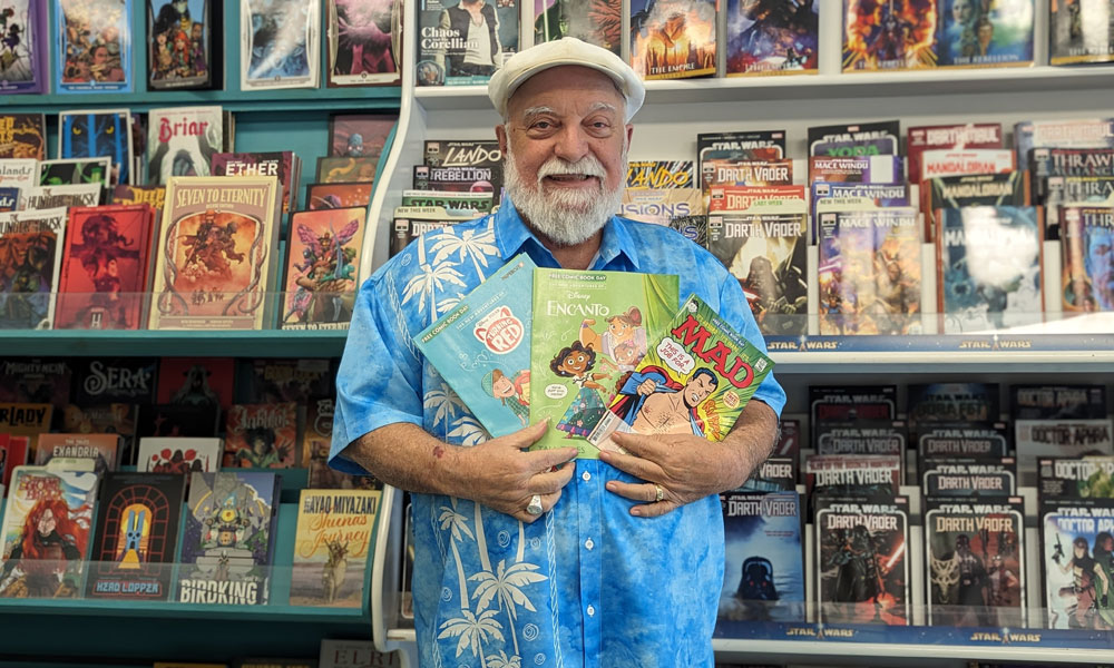 Man holding up three titles in front of a rack full of comic books