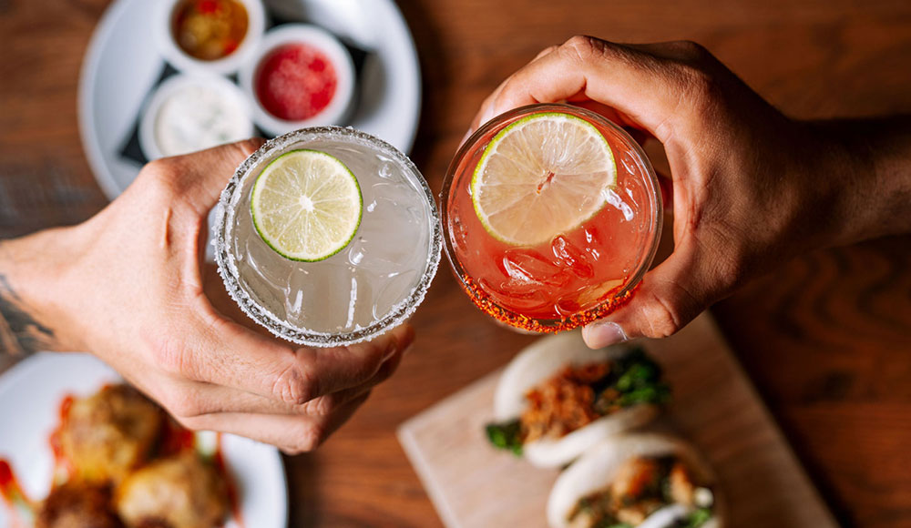 Two hands toast with cocktails over a table with various dishes