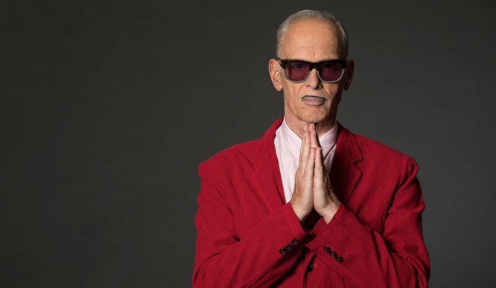 A John Waters Christmas Photo of man in a red suit wearing sunglasses and holding his hands as if praying