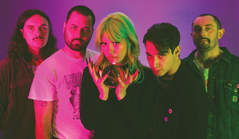 Band photo of one woman and four men illuminated with a pinkish light