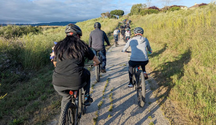 Two Nonprofits Keep Local Cyclists Rolling Along Group of cyclists riding on a trail through open space, seen from behind