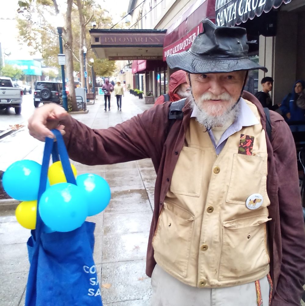Folk musician Dennis Holt standing on the sidewalk holding a bag with balloons.