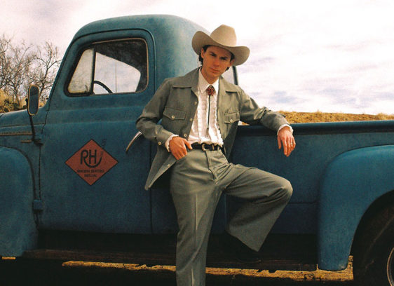 Ambient Wave Hayden Pedigo in a ten-gallon hat, white shirt with string-tie, trousers and coat, leans on a vintage pick-up truck