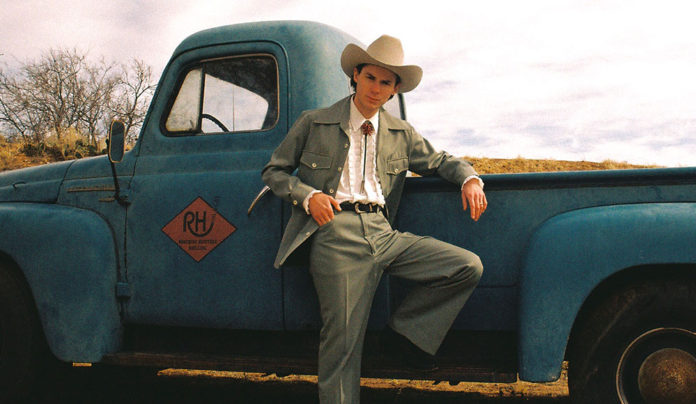 Ambient Wave Hayden Pedigo in a ten-gallon hat, white shirt with string-tie, trousers and coat, leans on a vintage pick-up truck