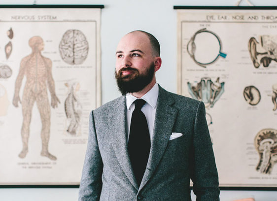 Keepin’ It Weird Jesse Thorn in buttoned gray suit, white dress shirt and long black tie stands in front of anatomical posters.