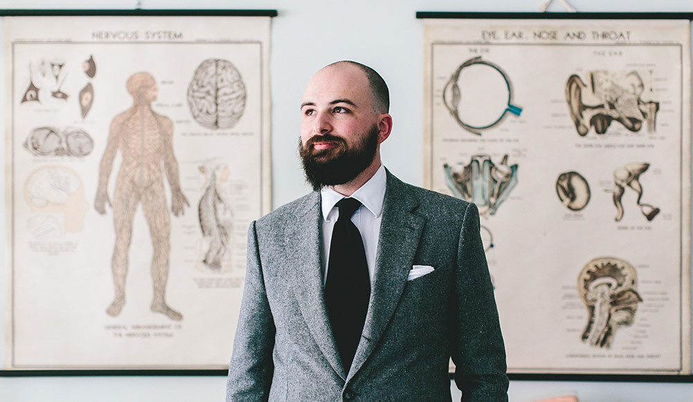 gtw2544_news1 Jesse Thorn in buttoned gray suit, white dress shirt and long black tie stands in front of anatomical posters.