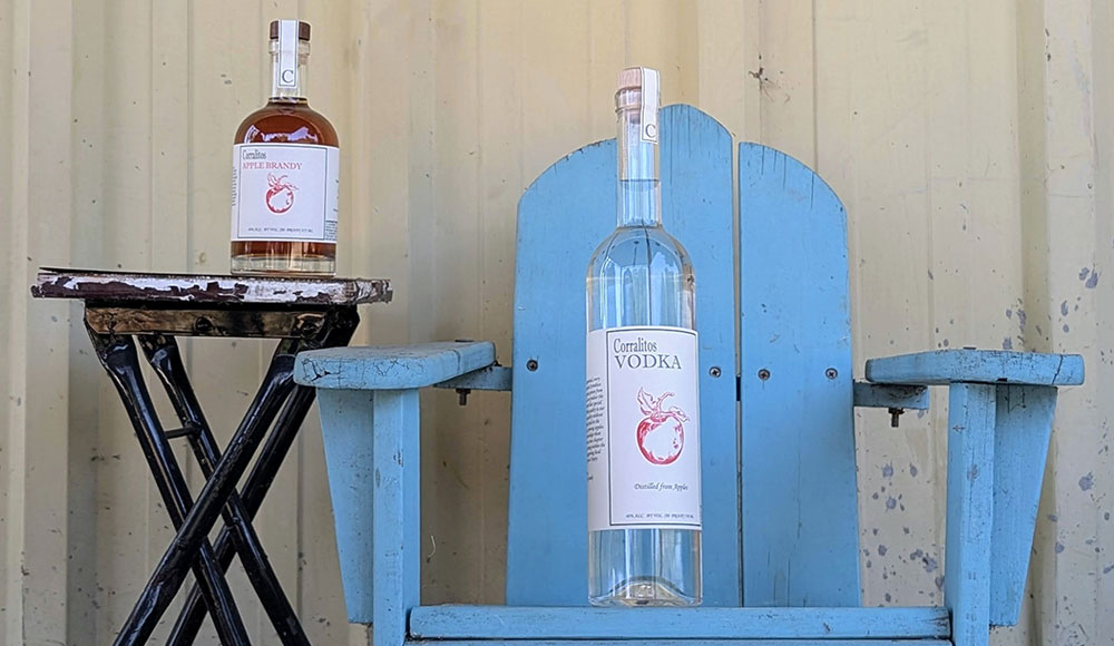 Bottles of Corralitos Apple Brandy and Corralitos Vodka displayed on a rustic blue chair and small table outdoors.