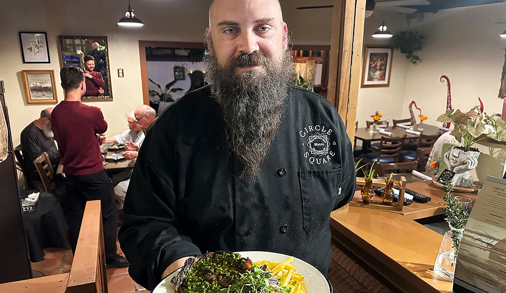 Chef Tyler Reiner holding a plated entrée inside a warmly lit restaurant dining room