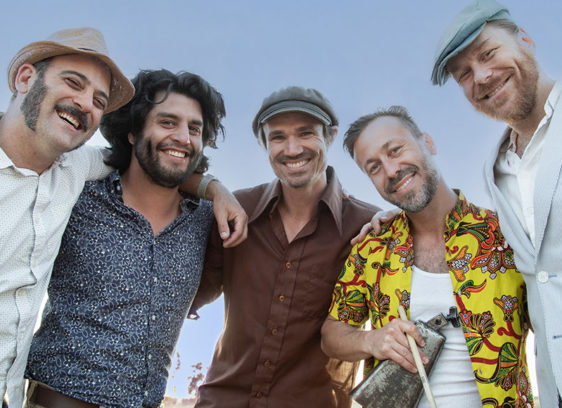 Digging Deep Roots The California Honeydrops band members stand together smiling outdoors, arms around each other.