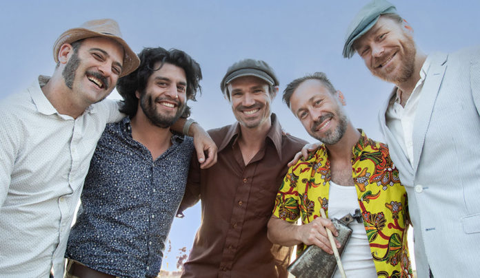 Digging Deep Roots The California Honeydrops band members stand together smiling outdoors, arms around each other.