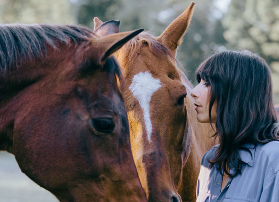 Nashville Abounds Singer Nicki Bluhm stands closely with two horses, touching foreheads in a peaceful outdoor setting.