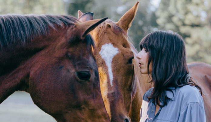 Nashville Abounds Singer Nicki Bluhm stands closely with two horses, touching foreheads in a peaceful outdoor setting.