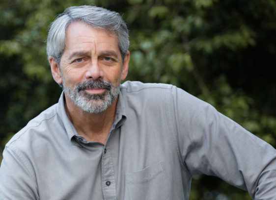 Being Here Now A gray-haired man with a trimmed beard and mustache poses outdoors in a light gray button-down shirt, looking calmly into the camera.
