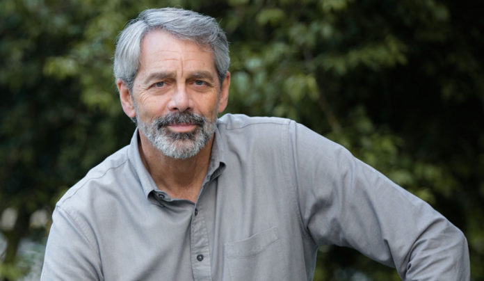 Being Here Now A gray-haired man with a trimmed beard and mustache poses outdoors in a light gray button-down shirt, looking calmly into the camera.