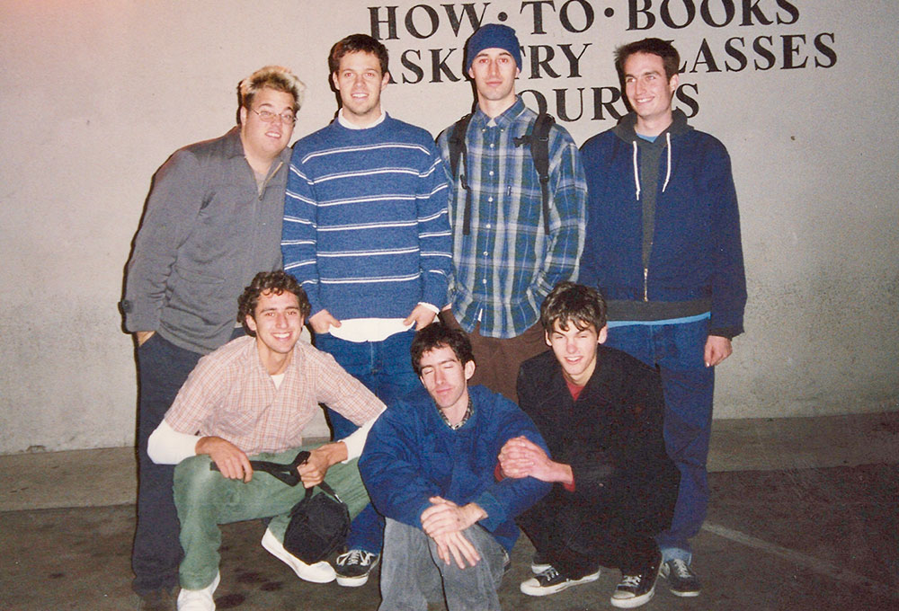 A group of seven young members of the band Slow Gherkin pose together against a wall, smiling and dressed in casual ’90s clothing.
