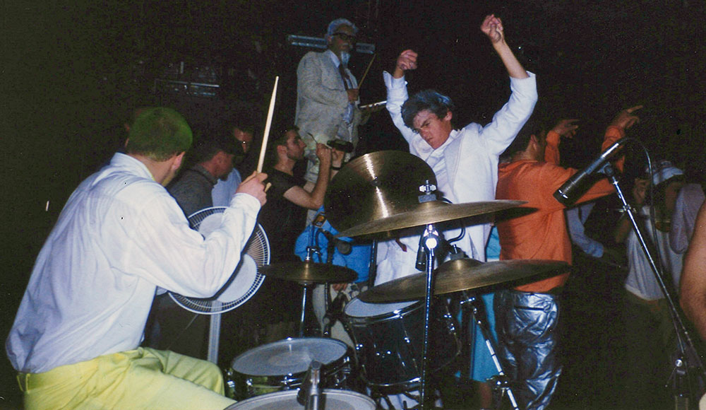 A drummer plays onstage as a costumed, energetic crowd surrounds him during an intense live Slow Gherkin performance.