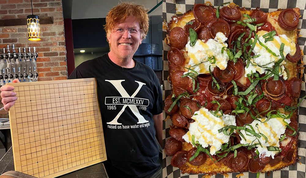 A man at d20 Pizza smiles and holds a wooden Go board beside a Detroit-style pepperoni pizza topped with ricotta and fresh basil.