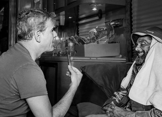 A man sits across from another man wrapped in towels, holding up a harmonica during an interview inside a dimly lit room or trailer.