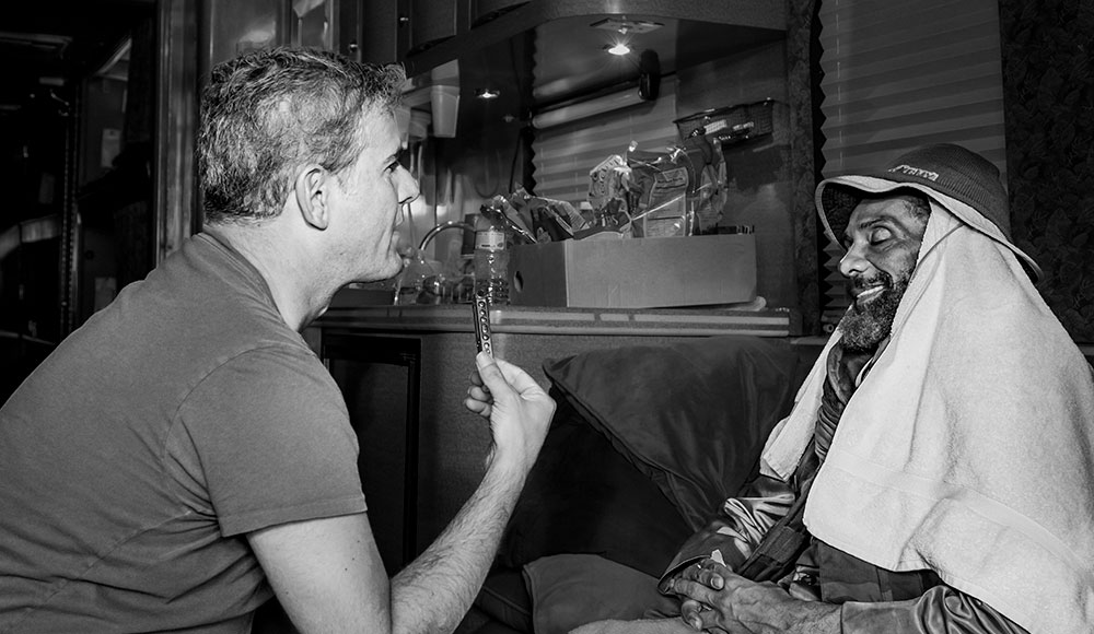 A man sits across from another man wrapped in towels, holding up a harmonica during an interview inside a dimly lit room or trailer.
