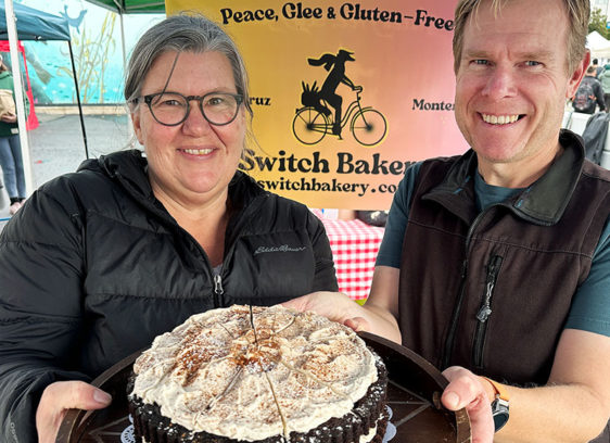 Switch It Up A smiling woman and man hold a chocolate layer cake with chai meringue at the Switch Bakery booth at a local farmers market.