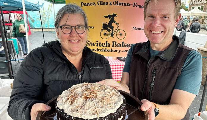 Switch It Up A smiling woman and man hold a chocolate layer cake with chai meringue at the Switch Bakery booth at a local farmers market.