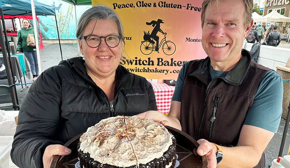 A smiling woman and man hold a chocolate layer cake with chai meringue at the Switch Bakery booth at a local farmers market.