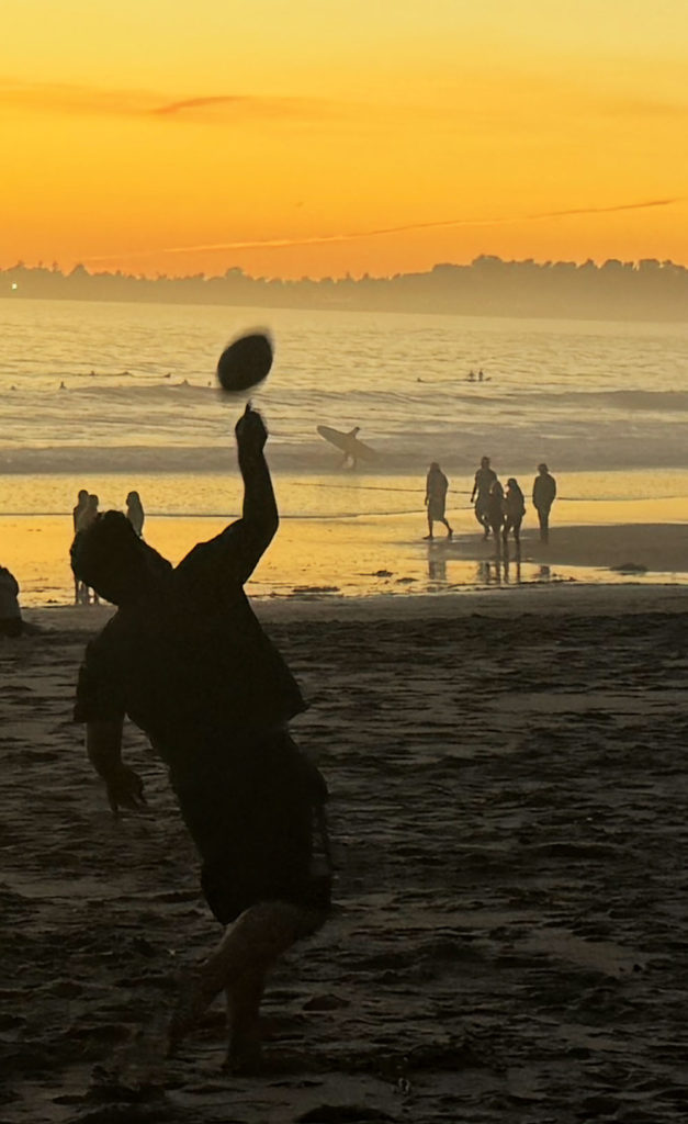 Silhouetted beachgoer throws a football at sunset as surfers and walkers move along the shoreline in the golden evening light.