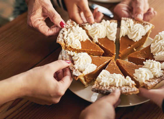 Healthy Holidays Hands from multiple people reach in to take slices of a pumpkin pie topped with whipped cream at a holiday gathering.