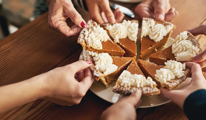 Healthy Holidays Hands from multiple people reach in to take slices of a pumpkin pie topped with whipped cream at a holiday gathering.