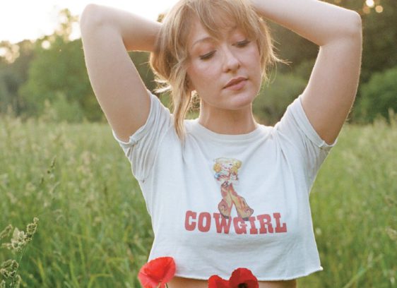 Things to do in Santa Cruz Singer-songwriter Alexa Rose standing in a sunlit field, wearing a “Cowgirl” crop top with arms raised behind her head.