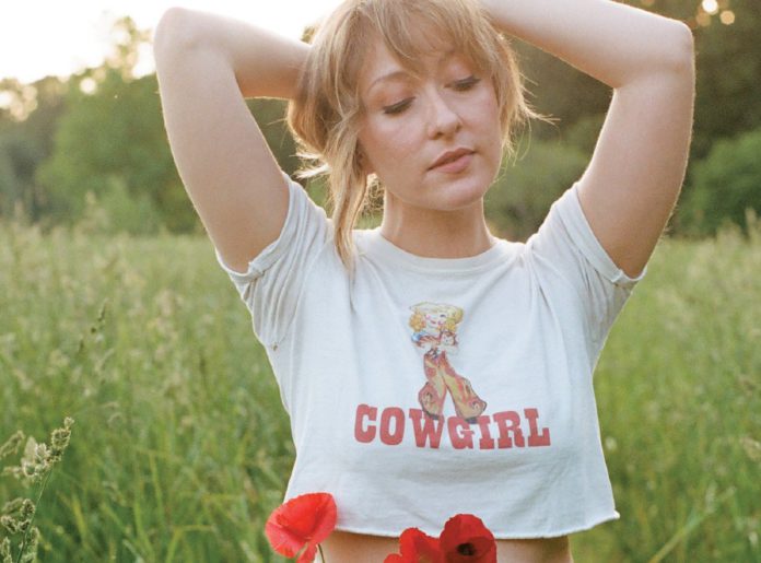 Things to do in Santa Cruz Singer-songwriter Alexa Rose standing in a sunlit field, wearing a โCowgirlโ crop top with arms raised behind her head.