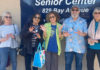 Hands-on Initiative A group of smiling seniors stand together outside the Mid-County Senior Center, holding up their hands playfully in front of the center’s sign.
