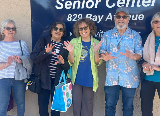 Hands-on Initiative A group of smiling seniors stand together outside the Mid-County Senior Center, holding up their hands playfully in front of the center’s sign.