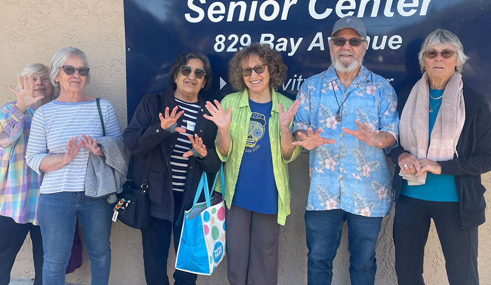 A group of smiling seniors stand together outside the Mid-County Senior Center, holding up their hands playfully in front of the center’s sign.