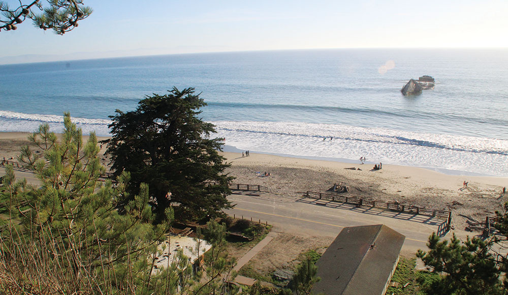 A high bluff overlooks Seacliff State Beach with waves rolling onto the sand and the remains of the S.S. Palo Alto visible offshore on a clear, sunny day.
