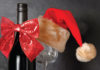 Shade of Red A wine bottle decorated with a red sequined bow stands beside a wine glass topped with a red Santa hat, creating a festive holiday scene.