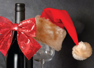 Shade of Red A wine bottle decorated with a red sequined bow stands beside a wine glass topped with a red Santa hat, creating a festive holiday scene.