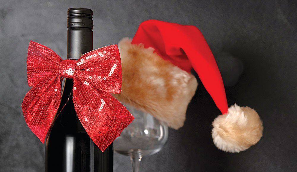 A wine bottle decorated with a red sequined bow stands beside a wine glass topped with a red Santa hat, creating a festive holiday scene.
