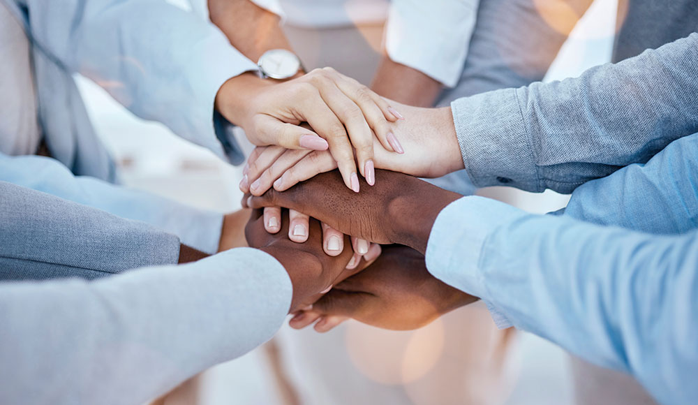 A diverse circle of people place their hands together in a stack, symbolizing unity, connection, and mutual support.