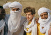 Things to do in Santa Cruz Four men stand shoulder to shoulder in bright desert clothing and white headwraps, smiling warmly in the sun against a textured tan wall.