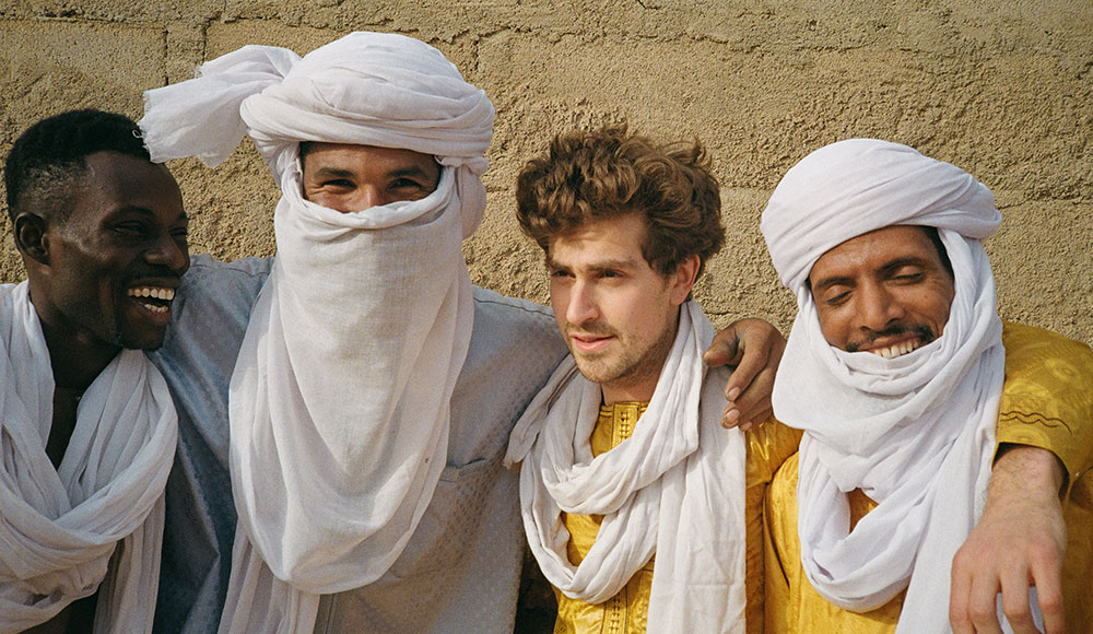 Four men stand shoulder to shoulder in bright desert clothing and white headwraps, smiling warmly in the sun against a textured tan wall.