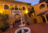 ‘Here’ Is Now A warmly lit Spanish-style courtyard with a central tiered fountain stands before the ornate arched entrance to Branciforte Plaza, featuring yellow stucco walls, balconies, and decorative columns at dusk.