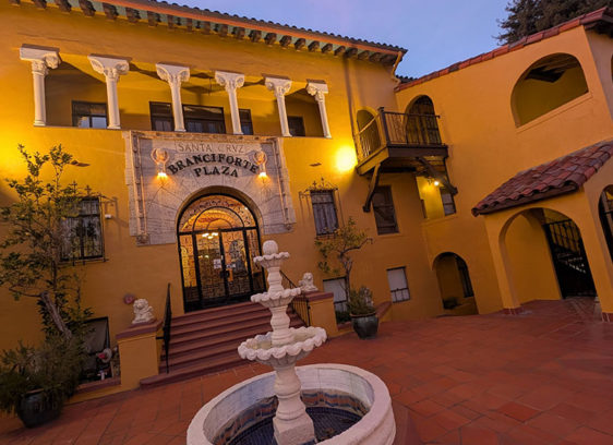 ‘Here’ Is Now A warmly lit Spanish-style courtyard with a central tiered fountain stands before the ornate arched entrance to Branciforte Plaza, featuring yellow stucco walls, balconies, and decorative columns at dusk.