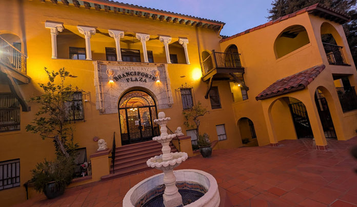 โHereโ Is Now A warmly lit Spanish-style courtyard with a central tiered fountain stands before the ornate arched entrance to Branciforte Plaza, featuring yellow stucco walls, balconies, and decorative columns at dusk.