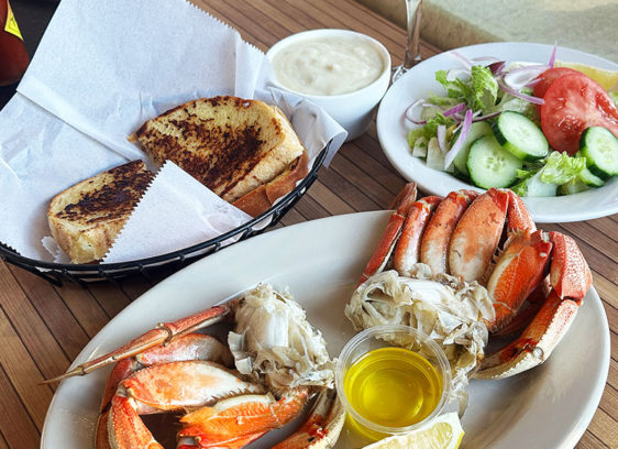 Ocean’s Bounty A plate of freshly cooked Dungeness crab served with melted butter sits beside bowls of clam chowder and a green salad, with toasted garlic bread in a basket on a wooden table.