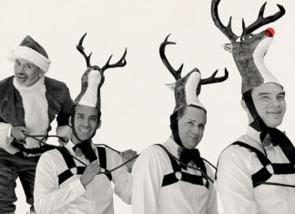 Mex-Mas Cheer El Vez dressed as Santa Claus playfully posing behind three performers wearing reindeer costumes with antler hats.