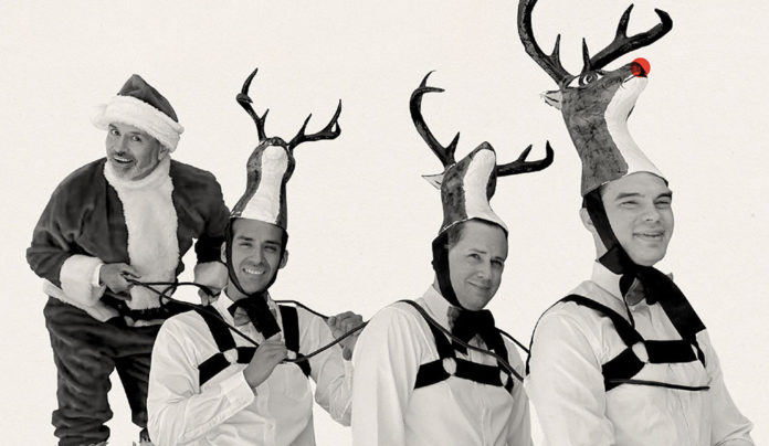 Mex-Mas Cheer El Vez dressed as Santa Claus playfully posing behind three performers wearing reindeer costumes with antler hats.