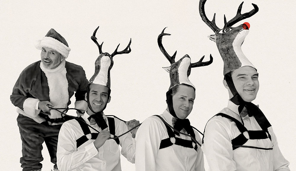 El Vez dressed as Santa Claus playfully posing behind three performers wearing reindeer costumes with antler hats.