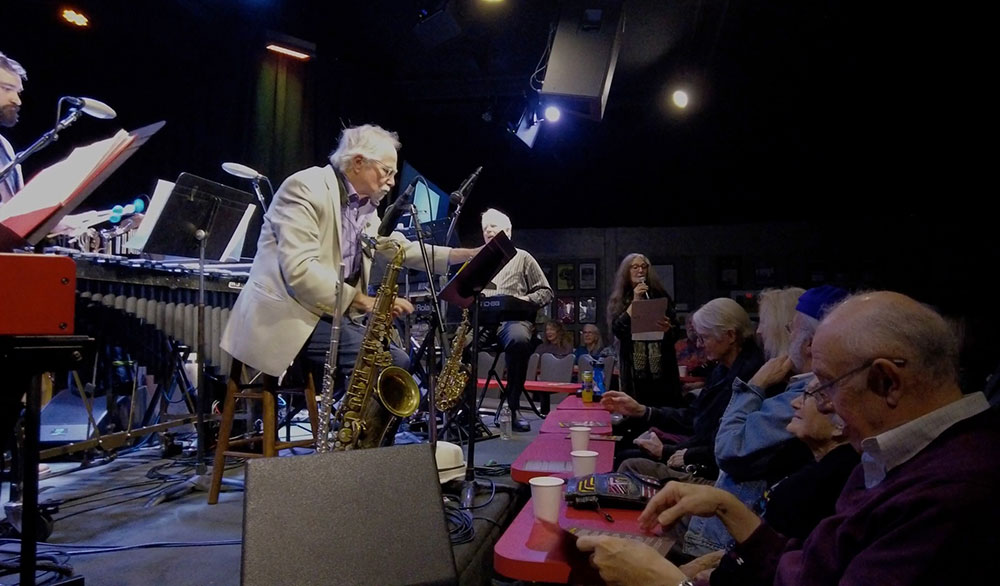 Musician on stage with horns surrounded by other players with the audience looking on